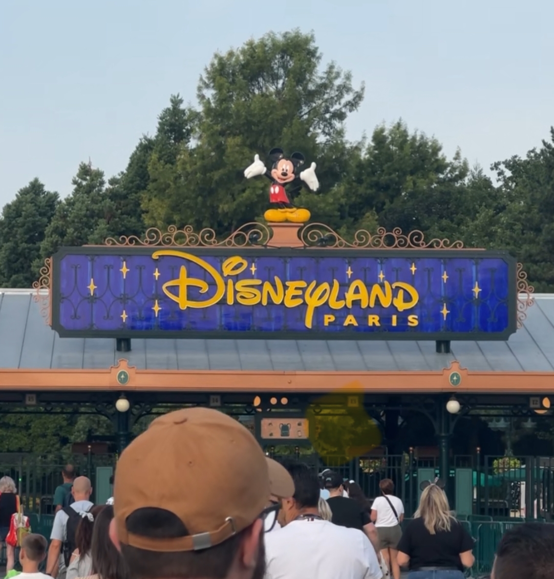 Mickey boven het blauwe ‘Disneyland Paris’-bord bij de ingang, met bezoekers bij de toegangspoorten